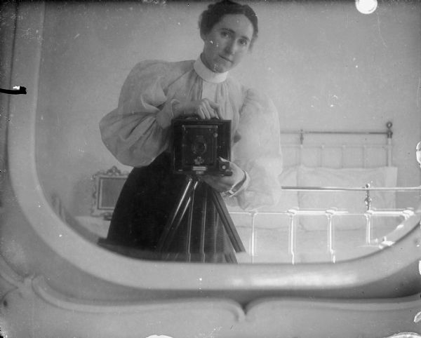 A female photographer takes a self-portrait in a mirror. The left and bottom frame of the mirror are in the foreground. Her camera is an Eastman Kodak Company Folding Kodet. The woman is dressed in a dark skirt and a light blouse with puffy sleeves and a high collar. Behind her is a shiny brass bed with pillows propped against the headboard, and on the left a framed picture stands on a nightstand.
