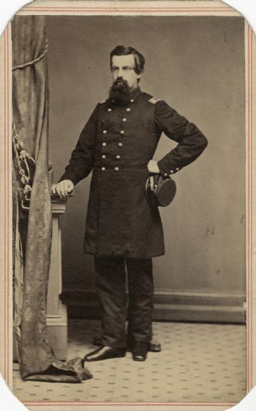 Colonel C.A. Wood posing for a full-length studio carte-de-visite portrait in his Union uniform. His right hand is resting on a plinth, and he is holding his hat in the other hand at his waist. He has a full beard and moustache. A drape with decorative cord partially obscures the plinth on the left.
