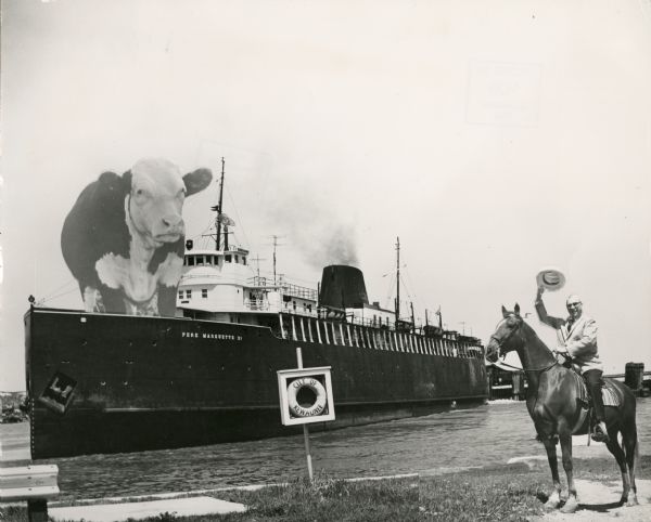 Composite photograph in the "Tall Tale" style. An enormous bovine stands on the front deck of the <i>Pere Marquette 21</i>, a steamer. On the right, on the shoreline, a man sits astride a horse waving his hat over his head. Nearby, a life ring inside a frame reads: "City of Kewaunee."