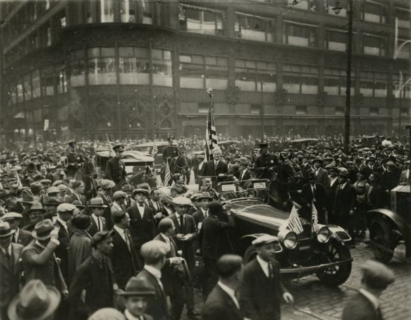 Presidential campaign photo of Robert M. La Follette, Sr. standing up in the back of a convertible automobile with an American flag behind him. Two signs in the windshield read "La Follette" and beneath, "Wheeler." More vehicles are turning the corner of State and Madison in a line coming from the right. His automobile is surrounded by a huge crowd of pedestrians. A police escort on horseback is providing security. In the background is a large multistory building with many windows and ornate decorations over the doors. To the right of the photograph in the scrapbook is written, "Let My People Go! Exodus 5-1." This photo was published by the <i>Chicago Tribune</i> with the caption "Chicago Admirers of Senator La Follette Greet Him On His Last Public Visit To City. This picture was taken at State and Madison streets in October of last year when the Wisconsin senator visited the city during the last weeks of the presidential campaign, speaking to big crowds."