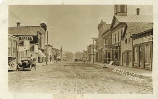 View down middle of unpaved street of the central business district. A man is sitting in the driver's seat of an automobile along the left curb near a garage. There is a sign for a livery further down the sidewalk. On the right is a shoe store, hardware store and doctor's office, and a sign in the shape of a clock. More automobiles are parked in the background, along with horse-drawn vehicles.