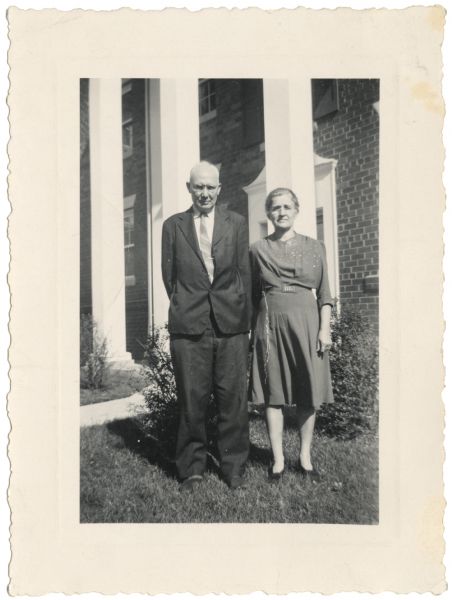 Full-length outdoor portrait of Hans and Clara Skott standing on the lawn in front of a brick building with white columns. Hans and Clara were the parents of Signe Skott Cooper.