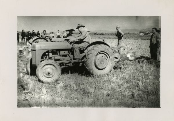 Hans E. Skott demonstrates use of a tractor outdoors in a field. A line of Chinese farmers watch in the background. Two other men stand on the right.