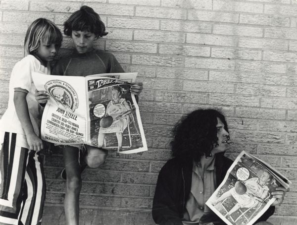 Individual crouches on the sidewalk against a brick wall in front of the Rennebohms at Lake and State Streets (now Walgreens) hawking <i>Take Over</i> newspapers.On the left, two boys stand leaning against the wall while reading a copy of the newspaper. Graffiti is barely visible in the upper right corner of the wall. On the front page of the paper is a photograph of a woman seated in a folding chair. The back cover is an advertisement for the [xxxxxx xxxxxx] 1972 Music Festival and Renaissance Art Fair. Featured bands are John Steele & Company, Grateful Dead, Taj Mahal, Country Joe, Ruby Jones, Commander Cody, Dr. Bop & Bungi. More local bands include Baby Grand, Circus, Tayles, Spring Green, Benedict, Phoenix, Ziggy & the Zeu, Blue Tailfly, Carrots, Moebius, Stark Weather Oil Slick, Speed-O-Meter, Pheet Phew, Punk-ola, Fat Sal and R.E. Taylor. The Festival would be held on August 18th, 19th and 20th.