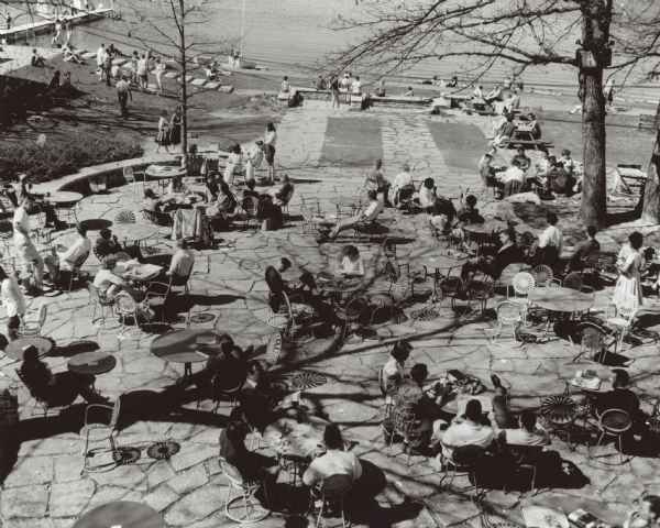 Elevated view of the flagstone terrace. People sit at tables in the  sunburst chairs. There are shadows of the empty chairs on the terrace as well as the shade of a tree. The Lake Mendota shoreline is in the background. There is a pier is in the upper left corner with several sailboats pulled up on the shore nearby. Several people recline on the beach.