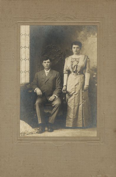 Full-length studio portrait in front of a painted backdrop of Beulah Irene Miller (1888-1958) and Charles Buy Thomas (1879-1925), a dairy farmer, posing in their wedding clothes.  The couple married February 21, 1912 in Evansville, Wisconsin.  

Beulah is standing on the right wearing a long dress, and Charles, wearing a suit, is seated in a chair on the left.