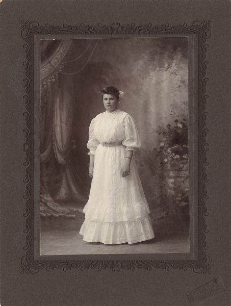 Full-length studio portrait in front of a painted backdrop of Beulah Irene Miller dressed for her graduation from Evansville (Wisconsin) High School. Her light-colored dress has a belt and a double ruffle on the bottom. She is wearing a bracelet on her arm and a bow in her hair.

Beulah was born January 18, 1888 in Evansville, Wisconsin, the daughter of Willis Exteen Miller (1859-1932), a farmer, and Sarah Maria Altemus (1857-1933).  She graduated from Evansville High School on June 7, 1906 in the evening.  The ceremony was held at the Evansville opera house.  Beulah was one of the eight students from the class of 30 graduates to give an oration at the graduation.  On February 21, 1912 Beulah married Charles Guy Thomas (1879-1925), a dairy farmer.  

