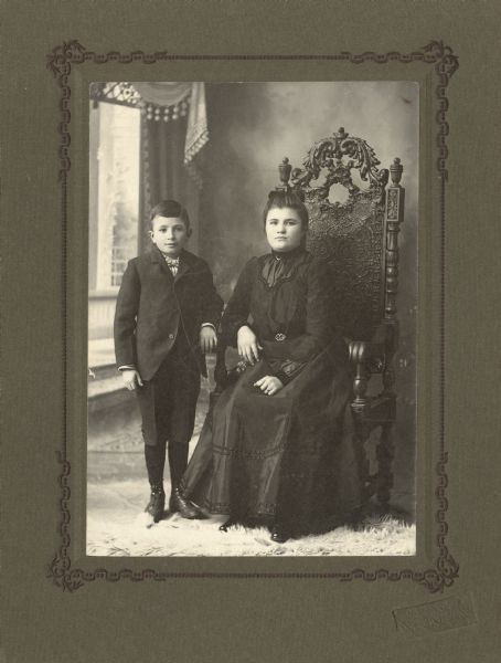 Full-length studio portrait in front of a painted backdrop of Beulah Irene Miller (1888-1958) and Willis Miller, Jr., sister and brother. Beulah is seated on the right in an ornate carved wood chair, and Willis is standing on the left with his hand on the chair arm. Beulah is wearing a dark dress with jewelry, and Willis is wearing a suit with knickers (knickerbockers), necktie, stockings and shoes. On the floor is a fur rug.

Beulah was born January 18, 1888 in Evansville, Wisconsin, the daughter of Willis Exteen Miller (1859-1932), a farmer, and Sarah Maria Altemus (1857-1933).  On February 21, 1912 Beulah married Charles Guy Thomas (1879-1925), a dairy farmer.