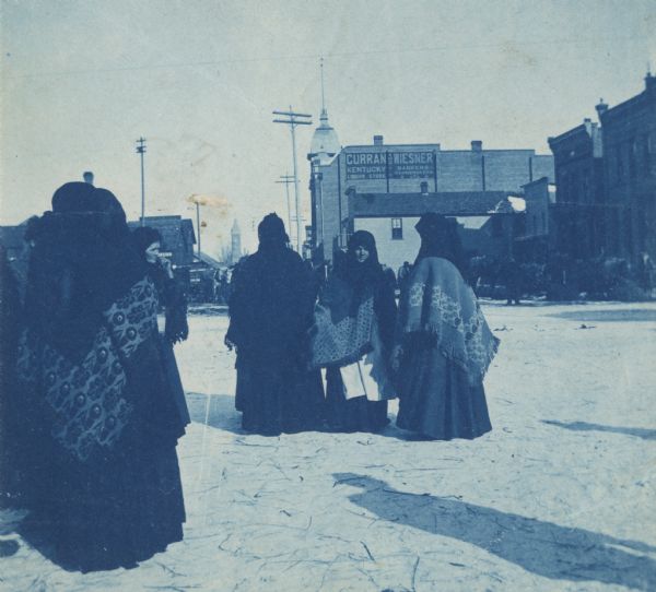 Polish immigrant women at the Market Square wearing traditional, national clothing. In the background are brick buildings. A sign on one building reads: "Curran and Wiesner. Kentucky Liquor Store. Bankers, Pawnbrokers."