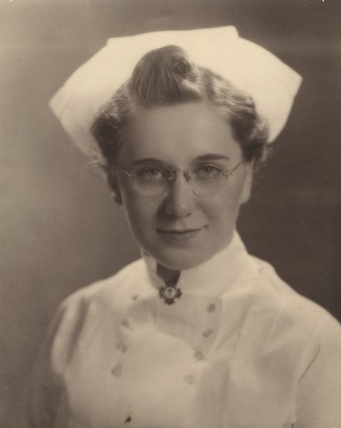 Quarter-length studio portrait of Fern L. Reinhardt. She is wearing her uniform, cap and eyeglasses, and a pin on the collar front displaying a cross, and was working at the U.W. Madison, University Hospital.