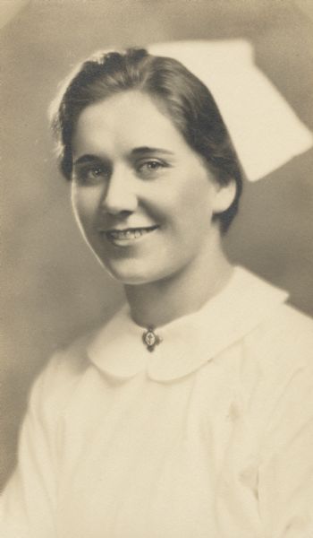 Quarter-length studio portrait of Fern L. Reinhardt, smiling on the occasion of her graduation from Nurse's Training. She is wearing her uniform and cap with a pin on the collar front displaying a cross.