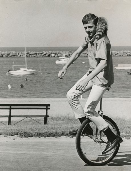Newspaper caption reads: "Passersby don't know it, but what appears to be a coonskin cap around the neck of Todd Vasas, 16, of 2773 S. Shore Drive, is the real thing. Todd, the live raccoon, and the unicycle attract attention as they ride along the lakefront. Todd is taking care of the animal for a friend who is on vacation." He is wearing a shirt, pants, socks and loafers. A rock breakwater and moored sailboats are in the background.