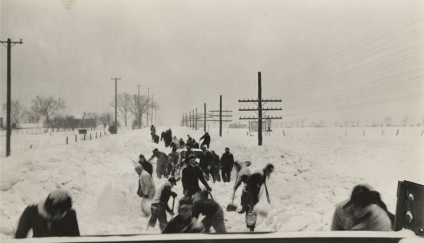 Slightly elevated winter scene with a large group of people digging a car out of the snow with shovels. The car can barely be seen in the center of the crowd. Power, telephone and telegraph poles line both sides of the road. Deep snow covers the ground.