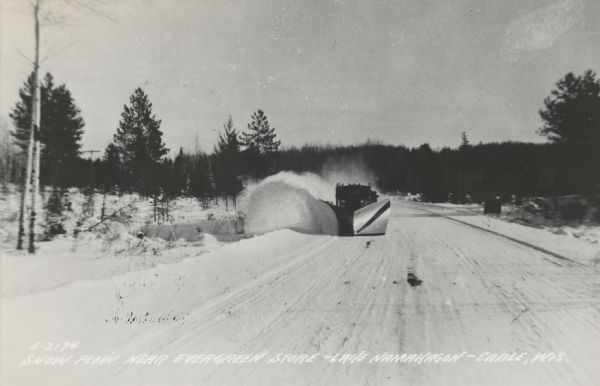 Winter scene with a V-blade snowplow from the front plowing snow from a road.  The snow is billowing to the left side of the road in a large arc. Trees are in the background.  Caption on foot of postcard, "Snow Scene Near Evergreen Store — Lake Namakagon — Cable, Wis."