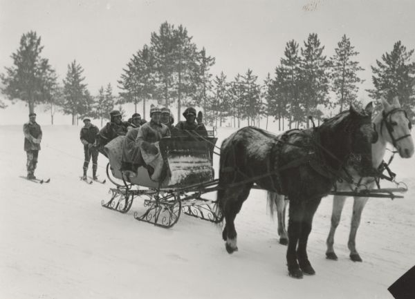 Winter scene with six well-bundled passengers in a sleigh pulled by two horses and pulling two skiers behind. In the background are pine trees.