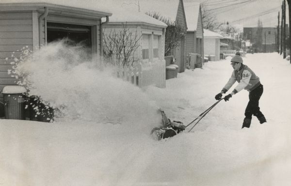 Winter scene with teenage boy snow blowing the driveway of a house. His letter jacket has "64" on the sleeve. He is also wearing a stocking cap with pom-pom, gloves and glasses. In the background is an alley with snowed-in garages, with trash cans and fences on the left, power poles on the right. A large building is in the far distance. Caption that ran with the photograph in the <i>Milwaukee Journal Sentinel</i>, "Michael Ridbert, 16, of 3808 N. 54th blvd., used a snow blower to clear a path to his family's garage."