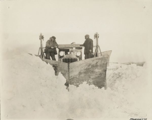 A winter scene showing two men standing behind the blade, on either side of the cab, of a Caterpillar Wausau-Plow Snow V-Plow. They are removing snow from the road.