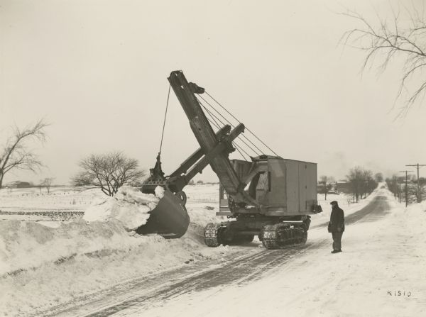 Winter scene with a gas powered shovel being used to move snow from a snow-covered road. A man is standing in the road to the right, watching. The shovel is on a Koehring crawler type excavator. In the distance are fields, fences, power poles and farm buildings.