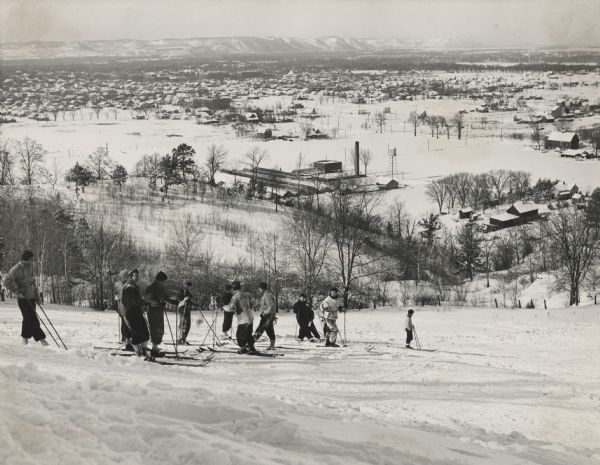 Winter scene looking down a hill towards skiers about to ski on the Capital Mississippi Alps near La Crosse, Wisconsin. La Crosse is in the background, and bluffs are in the far background.