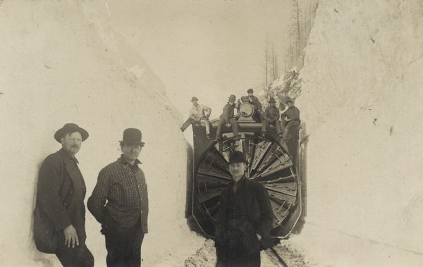 Winter scene with three men posing in front of a locomotive with a snow thrower attached to the front of the engine. The snow trough, barely wider than the locomotive, is as high as the train. Five men are posing on top of the engine. This was probably taken in Wisconsin during the big snowstorm of 1881. Between February 27th and March 4th, 1881, 2 to 4 feet fell in eastern and central Wisconsin with drifts up to 20 feet.