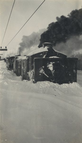 Winter scene featuring a Chicago, Milwaukee, St. Paul train from the front with snow thrower attached to the engine. Smoke billows to the right and power lines are on the left.