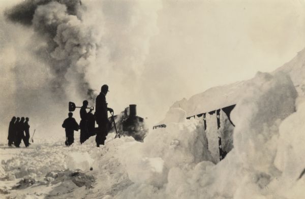 Winter scene with a group of men, many holding shovels, standing on the left watching a Chicago, Milwaukee, & St. Paul train pushing through snow, somewhere in northern Wisconsin. Smoke from the train billows high into the sky.