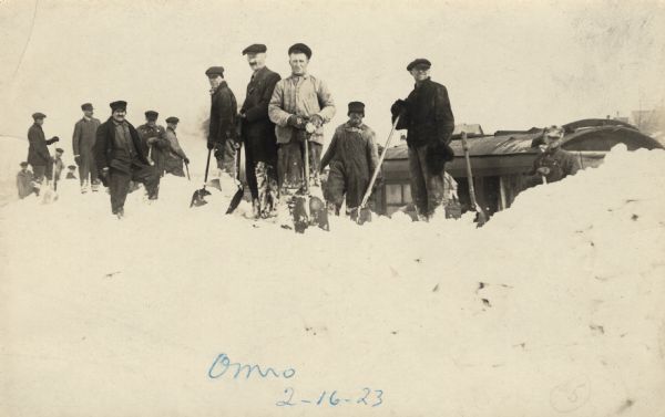 Winter scene with a group of men holding shovels pausing and looking toward the camera. Behind them is a railroad train in deep snow.