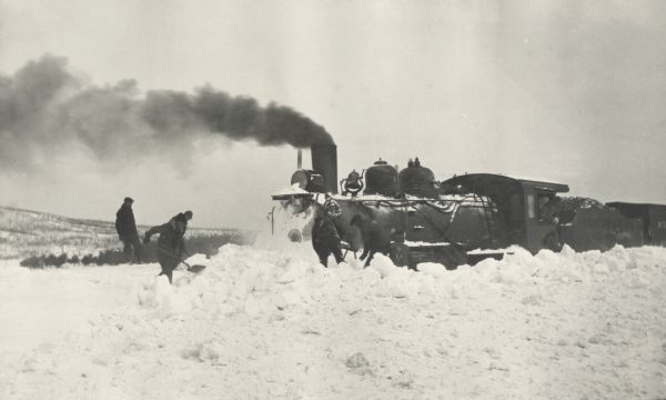 Winter scene with a Wisconsin & Michigan train pushing through snow. Men are shoveling snow in front of it. Smoke is billowing from the front of the train.