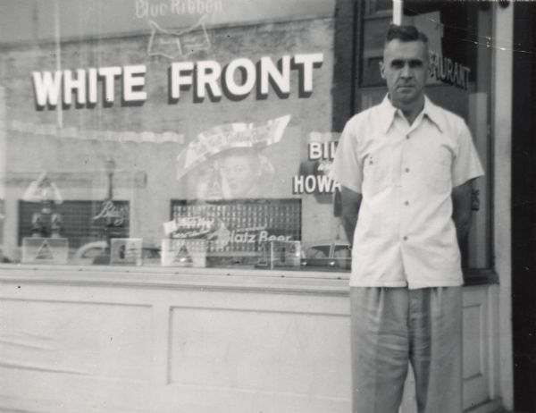 William R. "Bill" Kinney (1912-1978) stands in front of the White Front Tavern and Restaurant. Bill and his brother Howard Kinney owned the bar from 1948-1956. This photograph probably dates from 1950 or later, when the back room of the bar was converted to a small restaurant.