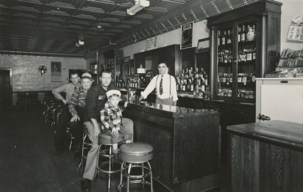 William R. "Bill" Kinney (1912-1978) behind the bar of the White Front Tavern. Bill and his brother Howard Kinney owned the bar from 1948-1956. Three unidentified men and a little boy, probably family members, are seated on stools at the bar. The boy is wearing a sailor's cap. The ceiling is pressed metal.