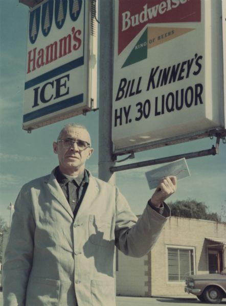 William R. "Bill" Kinney (1912-1978) stands in front of the electric sign for the Hi Way 30 Liquor Store, located at 3340 Commercial Avenue. Kinney owned the store from about 1961 until 1973. He is wearing a coat and eyeglasses and is holding up an envelope or document in his left hand. A brick building and an automobile are in the background.