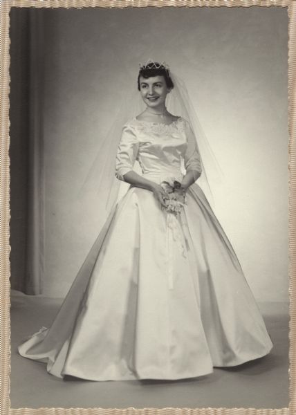 Formal full-length portrait of the bride, Mary (Kustermann) Esser (b. 1934). She wore a white satin princess-style gown with re-embroidered Alencon lace. She carried white orchids on a prayer book.