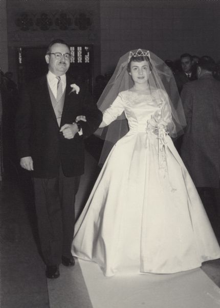 The bride, Mary (Kustermann) Esser (b. 1934) and her father, Alois F. Kustermann (1901-1993), walk down the aisle of St. Catherine Catholic Church. She wore a white satin princess-style gown with re-embroidered Alencon lace. She carried white orchids on a prayer book.
