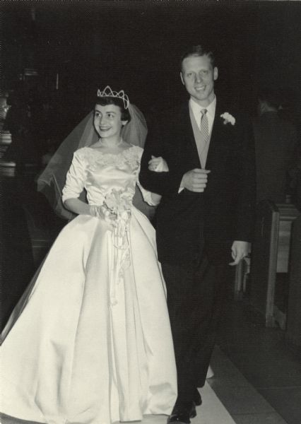 Mary (Kustermann) Esser and J. Thomas Esser smiling and walking back up the aisle after their marriage ceremony at St. Catherine Catholic Church. She is wearing a white satin princess-style gown with re-embroidered Alencon lace, and is carrying white orchids on a prayer book.