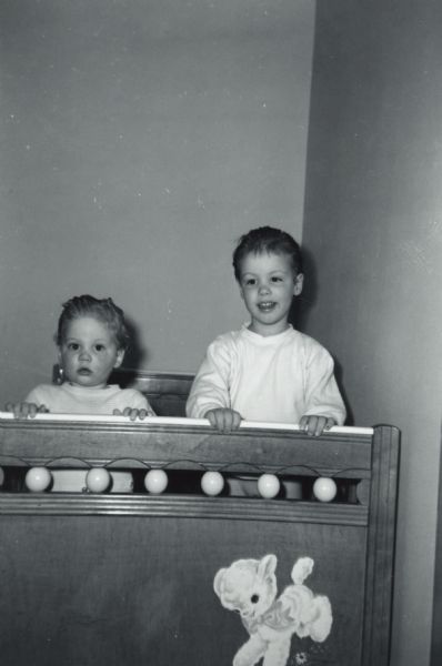The Esser sisters, Julie and Jane, stand in a crib with their hands on the foot board. A lamb decal decorates the crib.