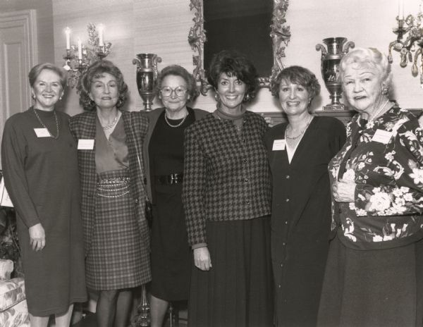 Group portrait of First Ladies of Wisconsin taken at the Executive Residence. Left to right: Sheila Coyle Earl; Dorothy Knowles Braun; Carrie Lee Dotson Nelson; Sue Ann Mashak Thompson; Elaine Thaney Schreiber; Mary Fowler Rennebohm. In the background urns are on a mantel or shelf, and an ornately framed mirror is in the center. Decorative lit sconces are on the wall.