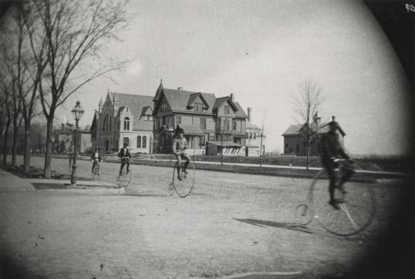 Five teenagers ride "High Wheeler or Penny-Farthing" bicycles down the street. Grand Avenue Congregational Church is in the background. Mature trees are on the left and newly planted trees are on the right.
