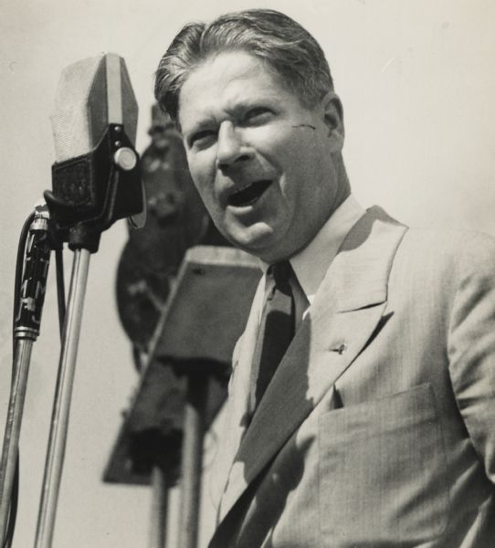 Philip Fox La Follette speaking into a WTMJ microphone. There are audio speakers behind him. He is wearing a suit and necktie.