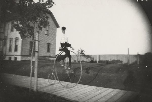 Young man riding a High Wheel or Penny-Farthing bicycle on a wooden sidewalk. There is a house behind the rider on the left. A young tree braced with a stake grows in the foreground. In the background is a wooden fence.