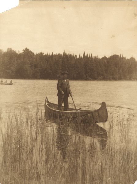 View across water of a Menominee man standing in a canoe in a marsh. He is holding a paddle in his hand. Behind him on the left is another canoe with three people. The far shoreline is lined with trees.