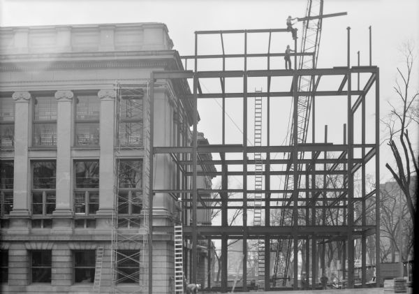 View from Langdon Street of steelworkers constructing the framework for the addition to the State Historical Society of Wisconsin Headquarters building. Park Street is on the right. Two workers add beams at the top of the structure with the help of a crane. More workers are on the ground below.
