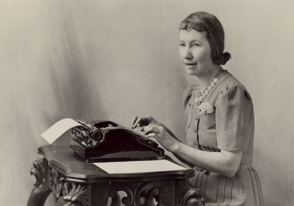 Louise Leighton poses sitting at her typewriter. She is wearing a dress, necklace, ring and an embroidered handkerchief in her pocket. The typewriter sits on an ornately carved table.