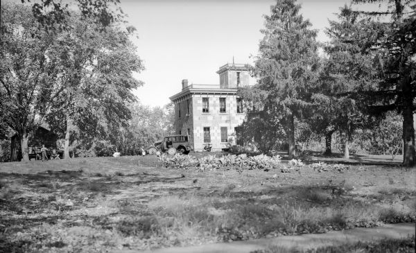View across lawn towards the T.C. Richmond House, which was located on the south side of Lakeside Street, between 505 and 605 W. Lakeside Street. The house is partially obscured by trees, and two men and two young children are sitting near an automobile in front. On the far left a young boy stands among trees in front of a small wood building.