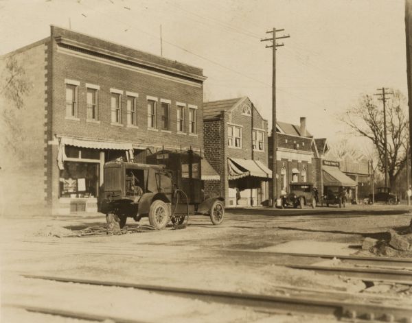 View across street looking west towards the north side of West Lakeside Street. Three known businesses are the South Side State Bank (3rd from the left), Universal Grocery Company (4th from the left) and Bontly's Arcade (5th from the left). The awning on the building on the left says "Hardware & Paint." A man is standing in the doorway of the building next to it. Two automobiles are parked at the curb. An Ingersoll-Rand truck with a compressor and jackhammers sits in the middle of the street. Railroad tracks are in the foreground.