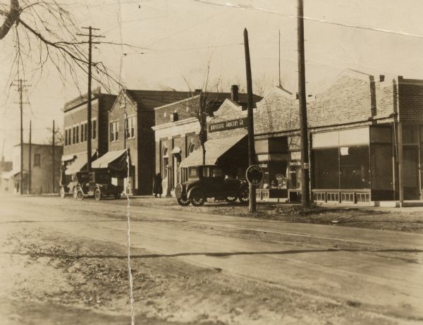 View across unpaved street towards the north side of West Lakeside Street, looking west. Three known businesses are the South Side State Bank (4th from the left), Universal Grocery Company (5th from the left) and Bontly's Arcade (6th from the left). The awning on the building (2nd from the left) reads "Hardware & Paint." The building on the right is for rent. Two women are walking into the bank, and three automobiles are parked at the curb. On the reverse is the text, "street needs paving, CT Dec. 31, 1925."



