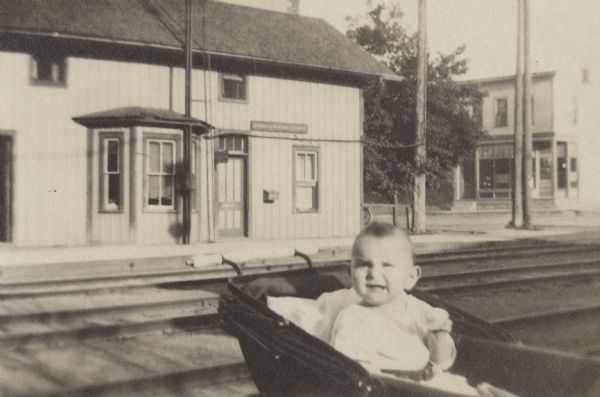 Exterior view of the South Madison Chicago and North Western Railroad Station located next to the railroad tracks that cross Lakeside Street. In the foreground is a baby sitting up in a baby carriage. In the background is a building that in 1931 would be the Kleinheinz Pharmacy.