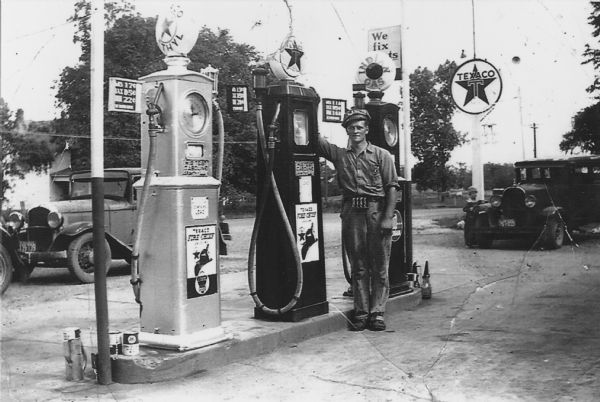 Lincoln Borgrud, in his work clothes, standing at the pumps at Droster's Grocery and Filling Station, 1438 Sherman Avenue. A child stands near a car beneath a Texaco sign in the background.