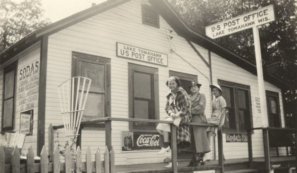 Exterior view of the Lake Tomahawk Post Office, with a woman and two young girls posed in front. A sign painted on the side of the building advertises "Sodas, fishing tackle, films-Developing, toilet articles, candy-cigars, ice cream."