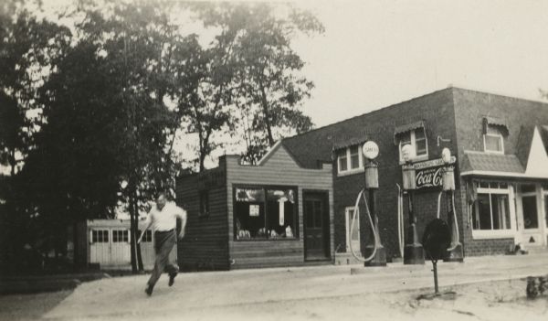 Exterior view of Droster Grocery & Fuel on Sherman Avenue. A man is running on the driveway in front, towards the camera. Trees are on the left.