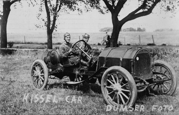 Two men, dressed in driving clothes, posed sitting in a Kissel car parked in a field. They are wearing caps, and their goggles are pulled up. In the background is a person riding a bicycle on a road lined with trees.